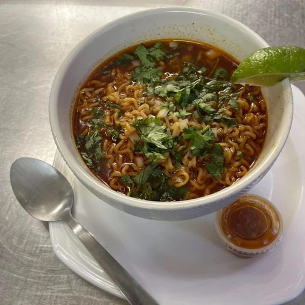 A bowl of Primos De Tico's Mexican ramen with cilantro, onions, and a lime wedge.
