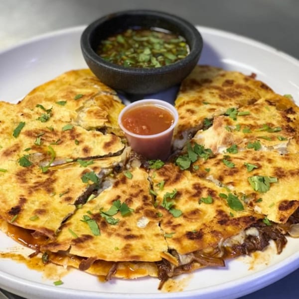 A large, golden-brown quesadilla cut into wedges, served with two dipping sauces on a white plate.